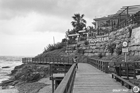 A woman walks along a wooden boardwalk near the sea, with a rocky shoreline and palm trees in view. The "Panorama Beach" sign is visible on a cliffside building under a cloudy sky.