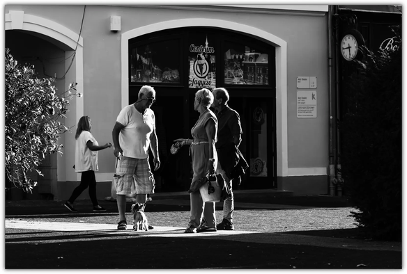 Ein schwarz‑weißes Straßenszenenfoto mit vier Personen und einem kleinen Hund: Drei Menschen stehen im Vordergrund vor einem Gebäude mit rundem Eingang und unterhalten sich, während eine weitere Person im Hintergrund vorbeigeht. Über dem Eingang hängt ein Schild mit der Aufschrift „Centrum Fagyizó“ Das harte Licht erzeugt starke Schatten und verleiht der Szene eine markante, zeitlose Stimmung.