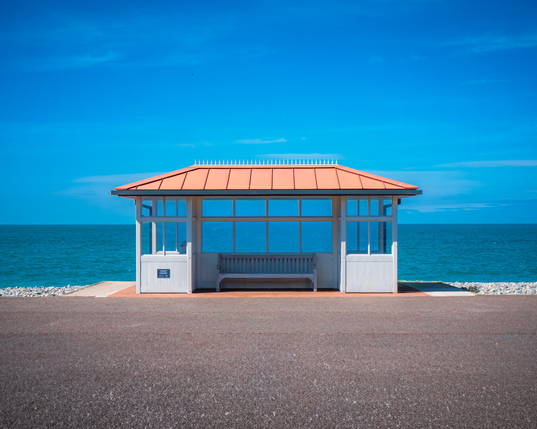 A symmetrical photo of a white Victorian seaside shelter with glass windows and a terracotta coloured roof. The structure is perfectly centred and stands on a paved promenade overlooking a turquoise sea under a light blue sky with wispy clouds.