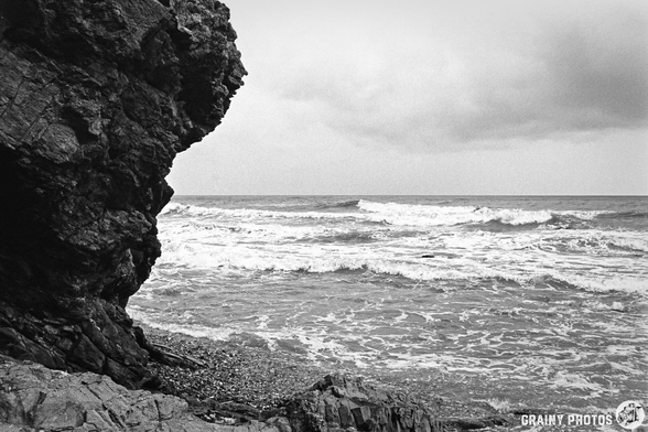 A dramatic coastline featuring a large rocky cliff on the left, with waves crashing against the shore under a cloudy sky. The image captures the raw beauty of nature in black and white.