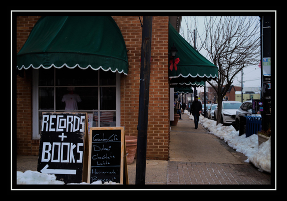A photo taken on a street corner, in color.
To the left of the image is a sign on the corner proclaiming that a record+book shop is located to the left of where the image is framed. Next to it is another advertisement for a garden cafe. Behind it is a dim shop window with a vintage dress on display. More on the right of the composition, a man walks up the sidewalk towards the viewer. There is some old snow piled on the edges of the sidewalks, and the day is overcast.