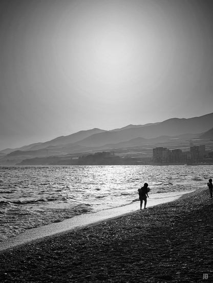 La imagen muestra una playa de guijarros en blanco y negro al atardecer o amanecer. El sol brilla alto en el cielo, iluminando suavemente el mar y creando reflejos sobre las olas. A la izquierda se ven montañas en capas que se difuminan en la distancia, y en la orilla hay dos siluetas humanas, una más cercana al agua y otra más alejada, caminando por la playa.