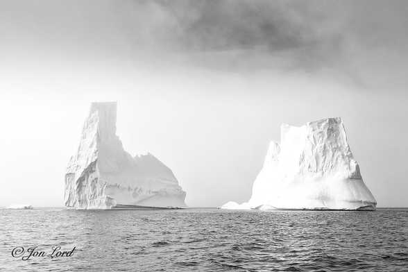 This is a black and white seascape photo in landscape format of a large iceberg with two tower-like structures at each end and a sea filled gap in the centre surrounded by low cloud and sea fog. Marguerite Bay, Antarctica (2024).

The ink black and gently rippled sea water rises from the base of the photo to a little less than the halfway up level where there is a crisp and straight horizon. The camera is about a metre above the water's surface and pointed directly at the iceberg that is about ten metres away. The iceberg, while single has the look of two icebergs close together. To the left of centre is the left half, having to look of a domestic clothes iron with a narrow, tall, tower like part at the blunt end of the iron with a small flat top. Most of the surface is very rough suggesting those parts have recently calved (large chunks have broken off to form new, smaller icebergs). Where the iceberg touches the water in the centre of the image (by the pointed end of the iron) there is a gap of a few metres with water freely flowing across it before the right side of the iceberg towers upwards. Remembering from school geography lessons that two-thirds or more of the iceberg will be submerged below the surface. The right part is larger than the left with no distinct shape other than a large triangle with the pointed top chopped off and with the similar rough surface to its friend on the left. Behind the iceberg is a mass of sea fog and low cloud, thicker on the left side of the photo and with the smallest break in the overcast above and to the right of centre. 

The location of this magnificent natural structure is Marguerite Bay, off the west coast of the Antarctic Peninsular, some two hundred km south of the Antarctic Circle.