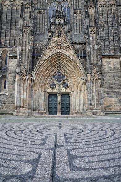Historical Magdeburg Cathedral’s ornate Gothic facade in dark and light stone, featuring intricate carvings, pointed arches, and tall stained glass windows between soaring spires and pinnacles. A large, multi-layered arched portal with decorative mouldings and sculptural details frames twin greenish bronze doors. In the expansive cobblestone plaza, a circular labyrinth maze pattern formed by alternating light and dark stones leads the eye towards the cathedral’s grand entrance.