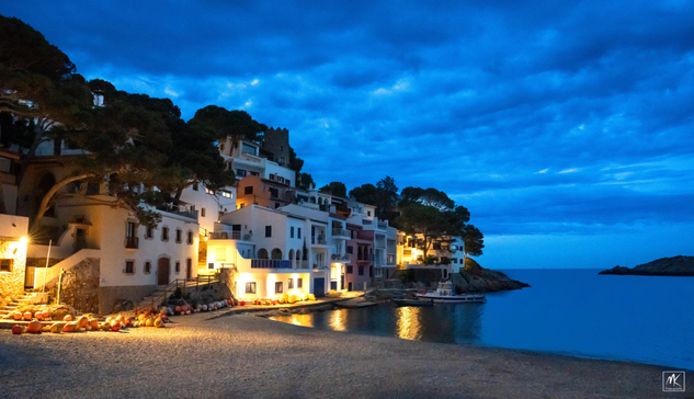 Color photo taken during twilight of the white buildings of a village on a cove on the Costa Brava, Spain that are partly lit by exterior lights. In the foreground is a pebbly beach and overhead is a dark blue cloudy sky. 