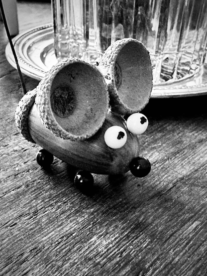 This black and white photo shows a self-made mouse made from an acorn. The fruit of the oak was used to depict the mouse's body, ears and tail. Small pins form the nose, eyes and legs of the mouse. It stands on a wooden table, behind it a drinking glass.