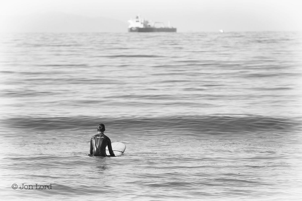 This is a black and white photo in landscape format of a lone surfer sitting astride his surfboard in a calm sea with an anchored cargo ship in the background. Sunset Beach, Orange County (2015).

Below and to the left of centre is the back of our lone, male surfer. He is viewed from the back with his back, the front of his board in view and at a distance of about ten metres. He is sitting astride his white board so that the front of the board is angled upwards at a slight angle. Our surfer has short, neat, dark hair: is wearing a black wetsuit and is looking straight ahead and out to sea. A short distance ahead of him is a small, about one metre high, rounded wave. There is not so much as a hint of surf anywhere in the photo. Theses small waves continue away from the camera. The medium grey/silver ocean continues upwards to just short of the top of the photo, where there is a poorly defined horizon. On the horizon and above the centre of the image is an anchored crude oil tanker, viewed obliquely with the stern and starboard (right) side in view, the bow angled to the right and at a distance of a few km. Above is a hazy sky, overexposed to white and devoid of any detail. 