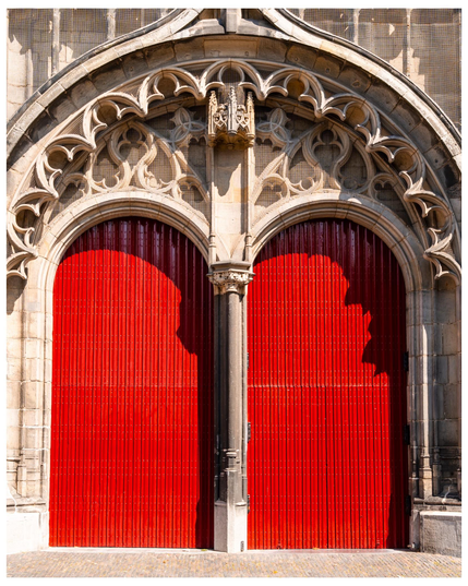 A grand entrance featuring ornate stonework and two large, vibrant red doors. The doors are adorned with vertical grooves and are set within an intricately designed archway. Sunlight highlights the architectural details of the stone facade.
