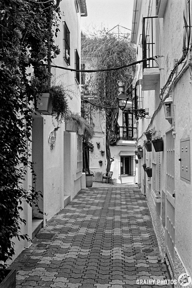 A narrow, cobblestone alley lined with whitewashed buildings, adorned with planters and greenery. Wires stretch across the path, leading to a glimpse of more structures ahead, creating a charming, serene atmosphere.