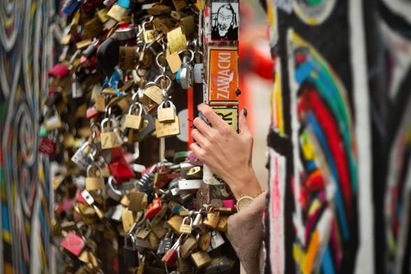 Farbfoto im Querformat. Zu sehen sind mehrere Stücke der Mauer in Berlin, sie sind mit Grafitti besprüht und mit vielen Liebesschlössern behängt. Zwischen zwei Mauerteilen greift eine weibliche Hand mit einer Armbanduhr und einer pelzigen Jacke durch, man sieht nur ein Stück des Armes, sonst nichts von der Frau. 