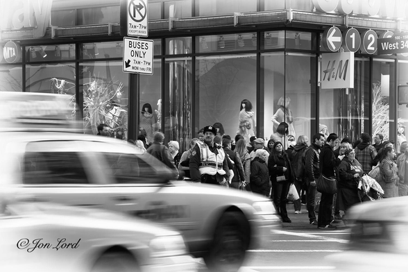 This is a black and white street photo in landscape format of a busy, city street corner with a traffic policeman directing traffic, blurred taxi cabs and a crowd of people waiting at the crossing. 5th Ave, NYC (2011).

In the lower left and right corners are the blurred images of New York taxi cabs speeding through the city's rush hour. The cab's are viewed obliquely with their right side and rear in view and from a distance of a few metres. In the centre of the photo is a New York Police Department traffic police man standing on the side of the road letting the traffic flow past. He is looking to his right (in the direction of the traffic) has a ushanka (Russian fur hat) on his head, is wearing wire rimmed glasses and has a dark moustache. He is wearing a sleeveless hi-vis jacket with 'NYPD' by the right shoulder and a thick, heavy winter's jacket beneath. Behind him is a crowd of commuters, shoppers and hardy tourists, all waiting to cross the street. Immediately behind is a large and brightly lit shop front belonging to the clothing chain H&M with numerous female mannequins behind the large glass shopfront. Above the centre and left of the photo is a street sign with an arrow and 'No Right Turn 7am-7pm except buses' and below a further sign 'Buses Only 7am-7pm Mon thro Fri'. In the upper right corner is a further street sign 'West 34th Street'.