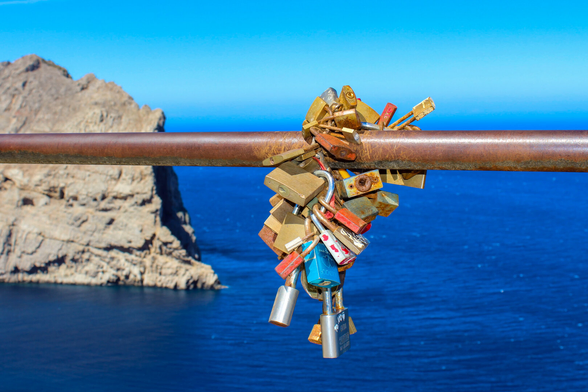 A close-up shot of a bunch of colourful padlocks locked together on a rusty metal railing. In the background, a rocky cliff by a deep blue sea under a clear sky.
