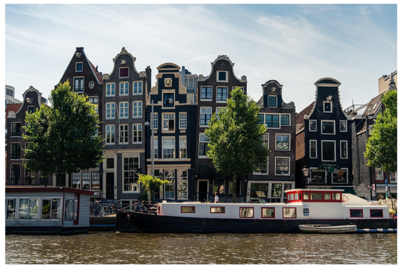 The image features a scenic view of traditional Dutch houses along a canal in Amsterdam, showcasing distinctive gable roofs. A houseboat with a red and white exterior is moored in the foreground, while lush green trees are positioned in front of the buildings.