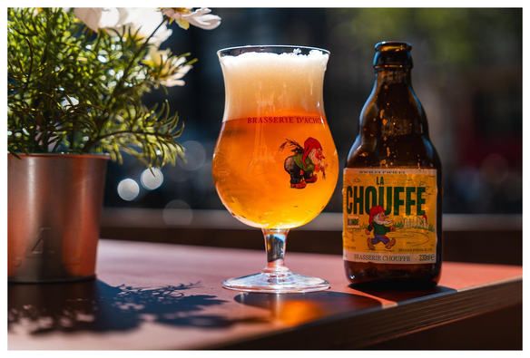 A glass of golden beer with a frothy top is displayed alongside a bottle labeled "La Chouffe." The glass features a whimsical gnome illustration. In the background, a potted plant adds greenery to the scene.
