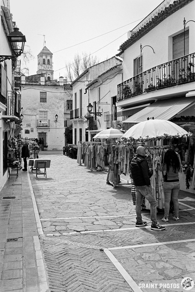A narrow cobblestone street lined with Souvenir Shops, featuring colourful clothing. Shoppers browse under white umbrellas, while a bell tower is visible in the background, framed by traditional Spanish architecture.