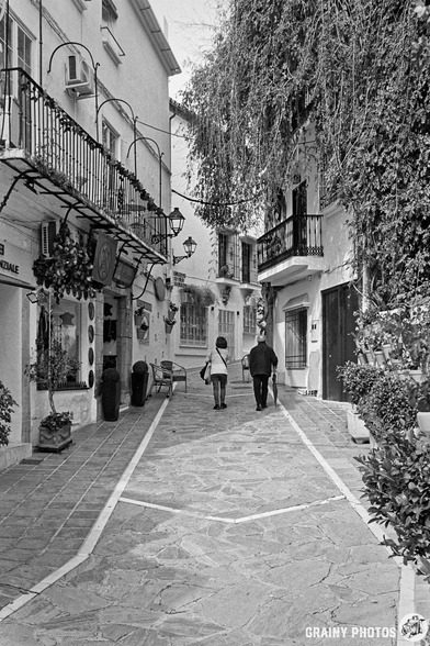 A couple strolls hand-in-hand down a narrow, cobbled street lined with traditional white buildings adorned with balconies and greenery, creating a charming, picturesque scene.