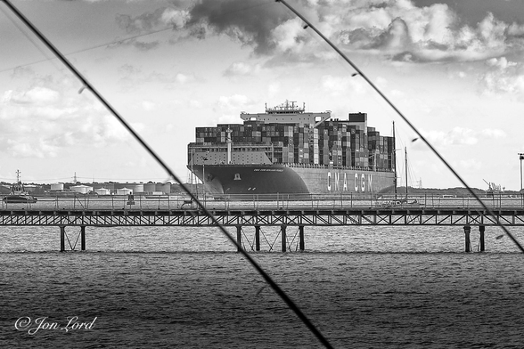 This is a black and white photo in landscape format of a giant container ship entering port and framed by two fishing rods and a metal pier. Hythe, UK. (2023)

The dark rippled harbour water rises from the base of the photo to the half way up level. Stretching diagonally across the image from the lower right are two fishing rods (fishing poles). One crosses below the centre of the photo and the other above the centre, both at an angle of about forty-five degrees. Below the centre and extending from left to right is a metal pier supported by a lattice metal frame and pillars driven into the seabed. In the centre of the photo and framed by the fishing rods and the metal pier is the bow of a large container ship, her hull extending away to the right underway from right to left. The ship is viewed obliquely with the bow and port (left) side in view and at a distance of about one km. The ship has a black hull with two white superstructures. One about a quarter of the way aft from the bow with the wheelhouse on top and the other about three-quarters of the way aft with a rectangular black funnel at the top. On the space forward, aft and between the superstructures are stacks of thousands of shipping containers, in places stacked nine containers high and twenty-one across, with more stowed below deck. Aft of the bow is the ship's name 'CMA CGM Benjamin Franklin' in white lettering with the shipping company's name in giant white lettering on the side of the ship in the centre 'CMA CGM'. On the far left of the photo is a single tug also underway from right to left and about to assist the container ship onto her berth in the port. Above is a partly cloudy sky with a few grey tinted, moody looking clouds. 

The ship is 399m long, 54m wide and dwarfed everything in the harbour. 