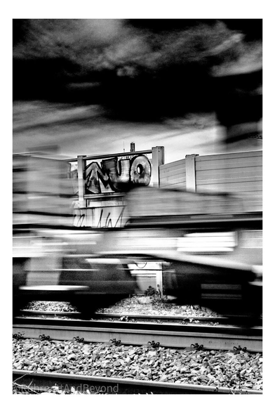 Eine Eisenbahnstrecke. Im Vordergrund ein durch Bewegungsunschärfe verschwommener Zuganhänger. Im Hintergrund ein Grafitto auf dem man deutlich ein "o" erkennt.