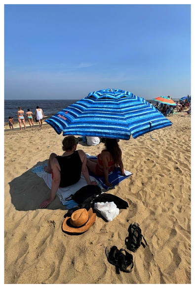 View of a beach on a sunny summer day, looking out to the sea. In the foreground, a couple sits on a blanket spread out on the sand, facing away from camera. They’re shaded by a large beach umbrella with horizontal blue and white stripes. At their backs are scattered belongings: black bags, a straw hat, a white sack, a pair of sandals. Farther away are more beachgoers, strolling along the surf or relaxing under colorful umbrellas. The sky is a clear blue, with vague clouds closer to the horizon. Taken at Sandy Hook Beach in New Jersey.