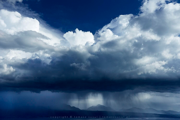 A dramatic landscape captures a vast expanse of towering cloud formations over a distant mountain range. The sky is filled with heavy cumulus clouds in shades of white and deep blue, creating a dense, textured canopy. Beneath these looming masses, distinct veils of rain fall toward the earth in steady vertical shafts. In the distance, the mountains appear as muted silhouettes against the hazy horizon. A thin layer of water at the bottom provides a still contrast to the intense activity above. This composition highlights the raw scale of an unfolding storm.