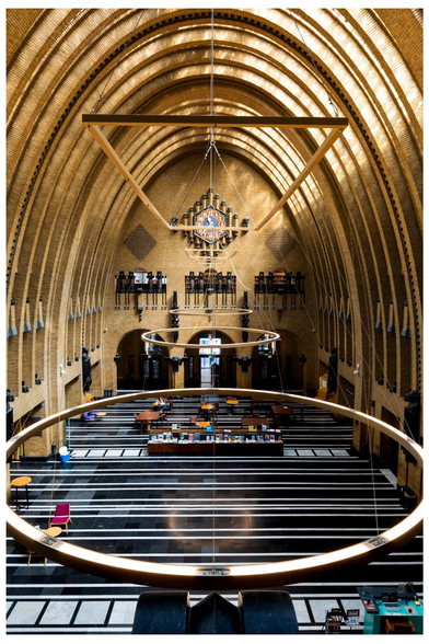 An interior view of a large, architecturally unique space featuring a high, arched ceiling with a warm color palette. Light fixtures hang from the ceiling, and the floor is patterned with contrasting black and white lines.