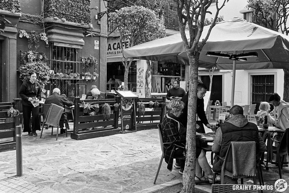 A busy outdoor café scene in Marbella Old Town  with diners enjoying meals at tables, surrounded by greenery and a vibrant atmosphere, while a musician plays nearby. The setting has a cozy, inviting vibe, typical of a social gathering spot.