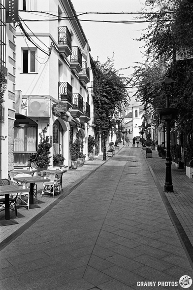 A quiet, narrow street lined with charming buildings featuring balconies and greenery. Tables and chairs are set outside cafés, while a few people stroll in the distance under soft light. The scene captures a serene urban atmosphere.