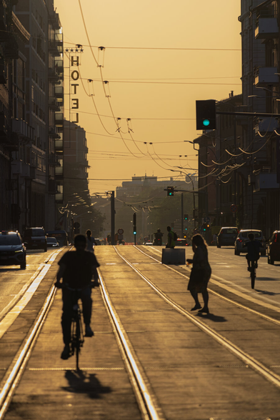 This is a photo of Via Saffi during golden hour. It is bathed in a warm, hazy orangey glow. We are  looking down a wide street with tracks that recede into the distance. There are several people which are silhouetted against the bright sky. Mainly a cyclist in the foreground and another cyclist and a pedestrian are a little further down the street. The traffic light up top on a pole to the right is green. Above there is a web of electrical wires for the buses which crisscross in the sky. On the left of the photo there is a tall building with a vertical "HOTEL" sign and stars above it. There are cars which are visible on the sides of the street.