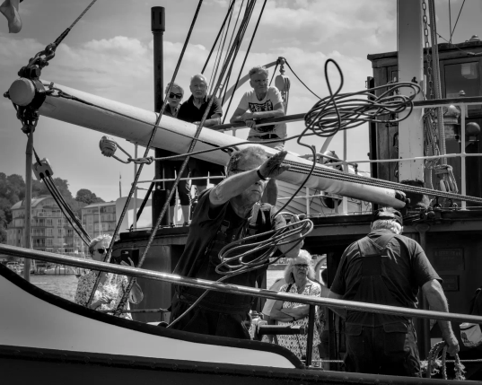 An Bord eines alten Dampfschiffes wirft ein Seemann die Festmacherleine über die Reling Richtung Pier. Viel Menschen stehen auf dem Schiff und schauen dem Seemann beim Anlegemanöver zu. 
.
On board an old steamboat, a sailor throws the heaving line over the rail towards the jetty. There are lots of people standing on the boat, watching the sailor as he moors the vessel. 
.
