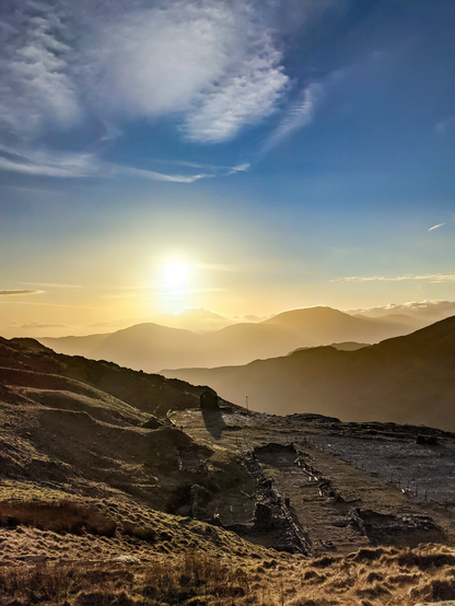 Silhouette of the historic stone ruins of an abandoned slate mining village on a mountain in Eryri National Park, North Wales. In the background a bright golden sunset shines through hazy ridges of distant peaks and a blue sky with light clouds.