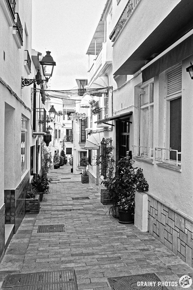 A narrow cobblestone street lined with whitewashed buildings, featuring wrought iron lanterns and potted plants. The scene captures a tranquil atmosphere in a quaint village alley, bathed in soft natural light.