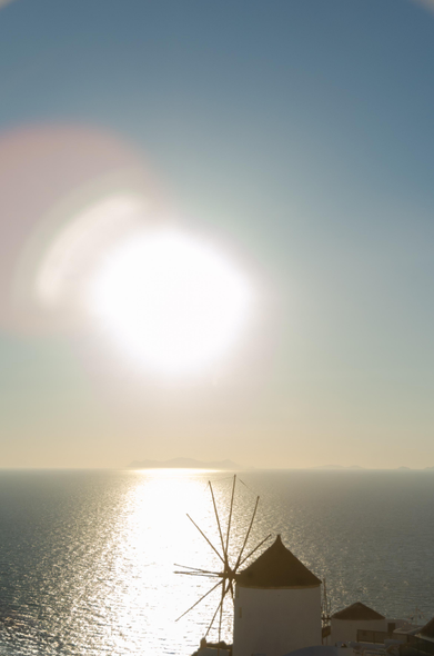 a photo I took in Santorini. it shows the ocean at sunset. the sun is in the middle of the frame, clear sky above, ocean underneath. a windmill or something at the bottom where the land begins. the sun leaves white over exposed reflection on the water and a little round flare around itself. 