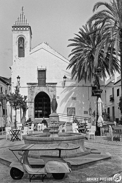 Ermita del Santo Cristo de Marbella in Plaza del Santo Cristo. A vintage scooter converted into a table stands beside a classic fountain, surrounded by other outdoor tables and chairs. In the background, palm trees create a tranquil, inviting atmosphere.