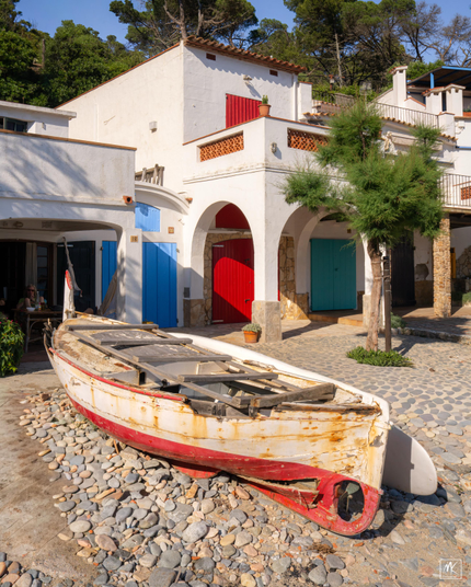 Color photo of a small rusted and dilapidated fishing boat on a pebbly beach in front of white stucco houses with brightly painted doors and shutters.
