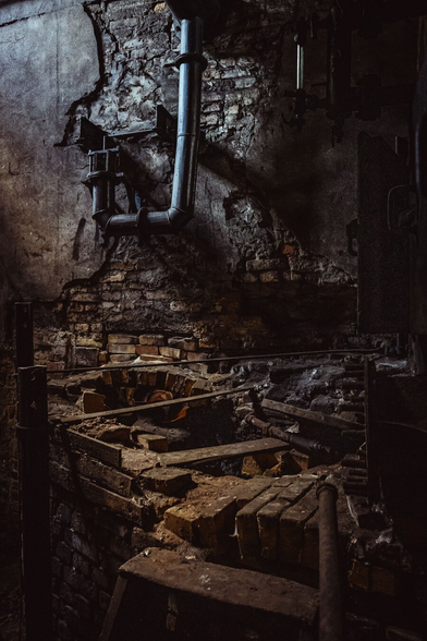 A vertical photograph showing a brick wall in an old iron forge. The wall is made of yellow bricks in different hues and is partially plastered, particularly in the upper left section. There are a number or dark metal pipes attached to the wall. In the bottom half of the image there is an opening in the wall and around the opening, a brick made channel comes out from the wall, open om the top, and a large pipe goes through the wall running inside the channel.

Everything in the image is old, worn and dirty, bearing witness to the heavy use of the forge through the years.