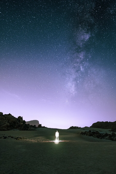 A person illuminated by a headlamp standing alone in a dark volcanic valley on Tenerife, Spain, dwarfed beneath a vast starry sky with the Milky Way stretching overhead.