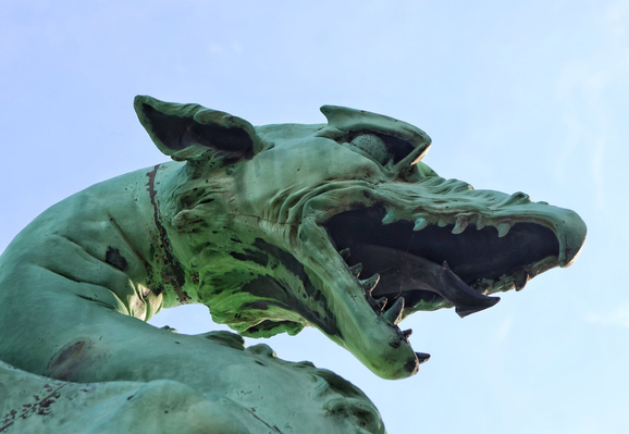 Close-up of a dragon statue from Ljubljana's Dragon Bridge, featuring a detailed copper sculpture with a weathered green patina. The dragon's mouth is open wide, revealing sharp teeth, a tongue, and a dark interior. The muscular neck is prominently visible against a pale blue sky.