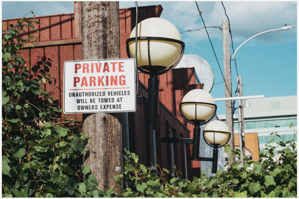 A "Private Parking" hangs on a wood utility pole in a parking lot. Green ivy can be seen growing at the bottom a red building with large round lights attached to it can be seen in the background.