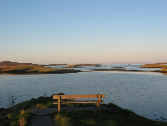 A picture of a bench seen from behind. Be Ch is sitting at the edge of water. Overlooking plenty of small islets with big island at the horizon. The Skye is blue with no clouds.