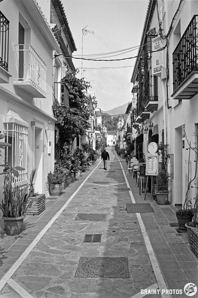 A narrow, cobblestone street lined with whitewashed buildings and pots of plants. A lone figure walks down the path, while balconies and signs add character to the quaint, peaceful scene. The black-and-white image evokes a timeless, charming atmosphere.
