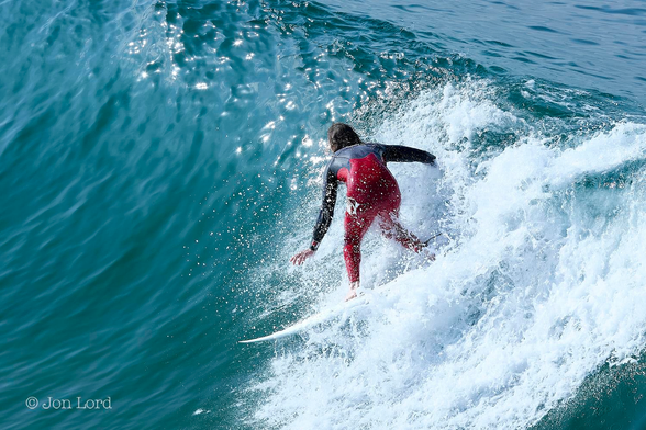 This is a colour water sports photo in landscape format of a sunlit surfer surfing down the front of a large breaking wave. Hermosa Beach, California (2014).

The entire image is filled with turquoise blue sea water lit by the still low morning sun. A large wave extends diagonally across the photo from the left to the right with the portion of the wave between the centre and right edge breaking and turning into a maelstrom of brilliant white foaming surf. In the centre of the photo is our male surfer. He is viewed from the side but with him standing sideways on his board and facing to the right so that his back is in view and at a distance of about five metres. He is surfing straight down the wave and in motion from right to left. He is wearing a colourful wetsuit that is bright red below the chest and dark blue above and down the long sleeved arms. His knees are bent as he crouches over his board with his arms outstretched as he maintains his balance. 