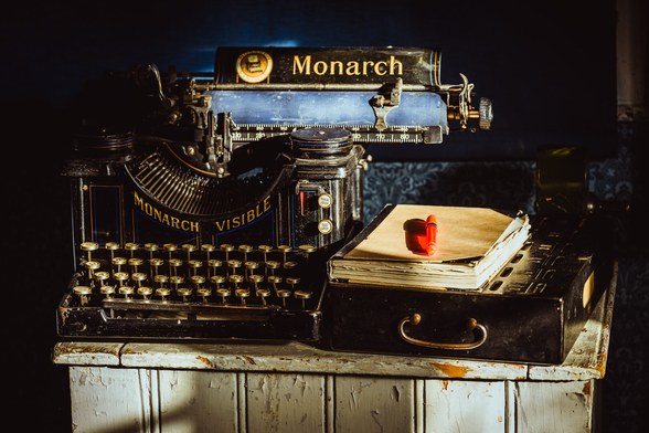 A photograph of an old typewriter, a black metal box and a worn notebook sitting on top of an old wooden cupboard, part lit from sunlight entering from the right through a windows outside of the photo, part deep in shadow.

The typewriter sits on the left side of the cupboard and is made of black painted metal, with brass details and  decorative blue and gold lines, which is quite tall. written on it is "MONARCH VISIBLE" with the lettering following a curve.

The carriage on top of the typewriter sticks out a bit to the right and has a blue roll, and a black metal plate above it that says "Monarch"

To the right of the typewriter a black metal box with brass handle sits on the cupboard, and a worn notebook with black back and cream white cover sits on top of the box. On top of the notebook lies a red plastic transparent pen.

The cupboard is made from white painted wood, quite worn and with the paint flaking off.

The wall behind is deep blue and in part with an intricate pattern.