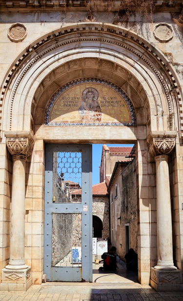 Ornate weathered light-coloured limestone archway entrance to the Euphrasian Basilica in Poreč, Croatia, adorned with intricate Romanesque floral and geometric carvings showing signs of age. Fluted Corinthian columns with elaborately decorated capitals flank the doorway, supporting a semi-circular tympanum with concentric stone arches and detailed mouldings. Above the metal lattice door, a golden mosaic with Latin inscription depicts religious figures in traditional Byzantine style. Two people are seated on the cobblestone pavement inside the archway, with historic stone buildings featuring red-tiled roofs and a clear blue sky visible beyond.