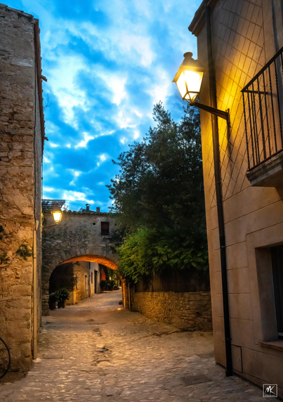 Color photo of an empty streetlamp-lit cobblestone street with an arched passageway in a medieval Catalonian town, taken after sunset with blue clouds in the sky above.