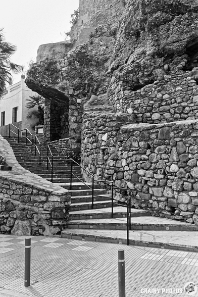 A stone staircase leads upward against the Marbella town wall. The scene is captured in black and white, highlighting the texture of the stones and the structure's rugged charm.