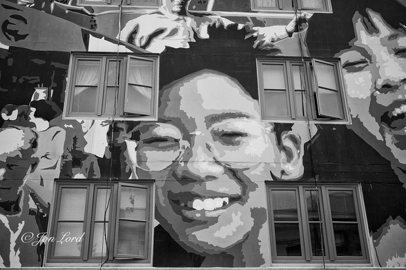 This is a black and white street photo in landscape format of the front of a building with a large mural of Chinese people painted on the facade. Chinatown, San Francisco. (2012).

The camera is facing the front facade of a city building, angled upwards slightly and viewed at a distance of a few metres (from the pavement). In the centre of the photo is the mural of a smiling young Chinese man, wearing glasses and looking towards the street and camera. In the four corners surrounding the centre: top left, bottom left etc, are four square windows made up of three rectangular, vertical panes of glass. Some of the windows are open, some closed, some with drapes, some with blinds: all very random. Above is the partial view of the base of the windows on an upper floor with just the base of the windows in view. To the right of the windows on the right side is a further large smiling face, I think a young Chinese woman. To the left of the left windows are more murals of Chinese people in an urban setting. 


