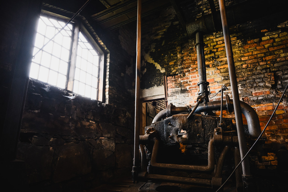 A photograph taken inside an old iron forge, facing a corner. On the wall on the left side are two large multi pane windows, slanted at the top, letting in bright light from the outside. The bight light makes the outside entirely invisible as the window panes are overexposed, creating a bit of blooming around the windows.

Along the other wall is a heavily used and worn Lancashire hearth, which was used to refine pig iron into wrought iron.

The walls are made of yellow and red brick, very dirty, worn and blackened after many years of heavy use.

In the corner is a door opening into another room.