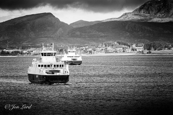 This is a grungy black and white photo in landscape format of two medium sized Norwegian car ferries passing each other in the Trondheimsfjorden, Norway (2023).

The dark and rippled waters of the fjord rise from the base of the photo to the halfway up level. To the left of centre is the first and closest ferry. She is viewed obliquely with the bow and starboard (right) side in view and occupying about a quarter of the length of the photo and at a distance of about twenty metres. The ferry is underway towards the camera and moving diagonally across the image. She has a black hull rising up about three metres above the waterline with two white superstructures on either side of the central car deck and a further white structure (passenger cabin) spanning over the car deck. On top of the passenger cabin is a large, control tower like, wheelhouse with large darkened windows and a forest of aerials on the roof. The vessel has a ramp on the bow, now raised, that is lowered when moored, to enable the cars, lorries and foot passengers to disembark. There is a further ramp on the stern where the operation is reversed, hence the name RoRo Ferry (roll on - roll off) so there is really no bow or stern, the ferry just goes forwards one way and reverse the other. There is a small white wake trailing behind the ship. In the centre of the photo is the other ferry, perhaps fifty metres away and identical to the first ferry but underway in the opposite direction, which is obvious with the white wake trailing behind the vessel. Here the stern is in view, which is identical to the bow of the closer ferry. This ferry is more distant having just passed the first ferry and steaming in an opposite direction. Beyond the ferries is the northern shore of the Trondheim Fjord with scattered low-rise buildings dotted along the coastline and with a tree line terrain rising behind with a few rolling hills on the left and a small rocky mountain on the right. Above is a moody, partly cloudy sky. 

The two ferries are the sixty-six metre long, MF Austrått and MF Vestrått that cross the fjord between Brekstad - Valset and back.