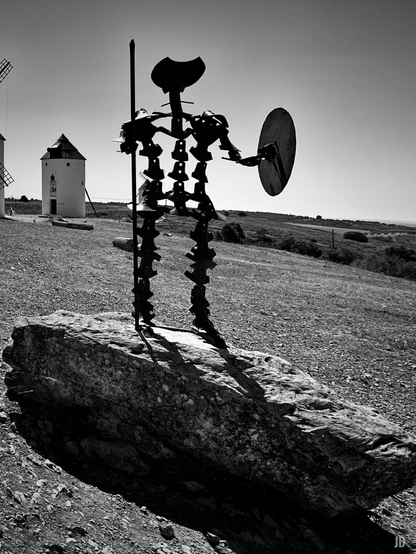 La imagen en blanco y negro muestra una escultura metálica que representa a Don Quijote con lanza y escudo, de pie sobre una gran roca. Al fondo, en medio de un paisaje abierto, se observan varios molinos de viento tradicionales sobre una llanura despejada.