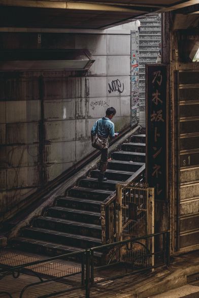 A man in blue with bag in stride, is climbing stairs where shadows hide. Beside the sign of timber black, he walks along the narrow track, while textures grey and markings small, are scattered on the concrete wall. A step by step to light above, he moves within the town we love.